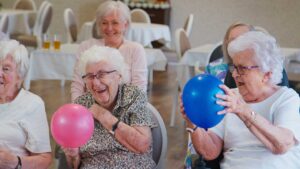 Older adults share a conversation and activity around a table in a bright assisted living common room.