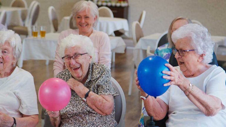 Older adults share a conversation and activity around a table in a bright assisted living common room.